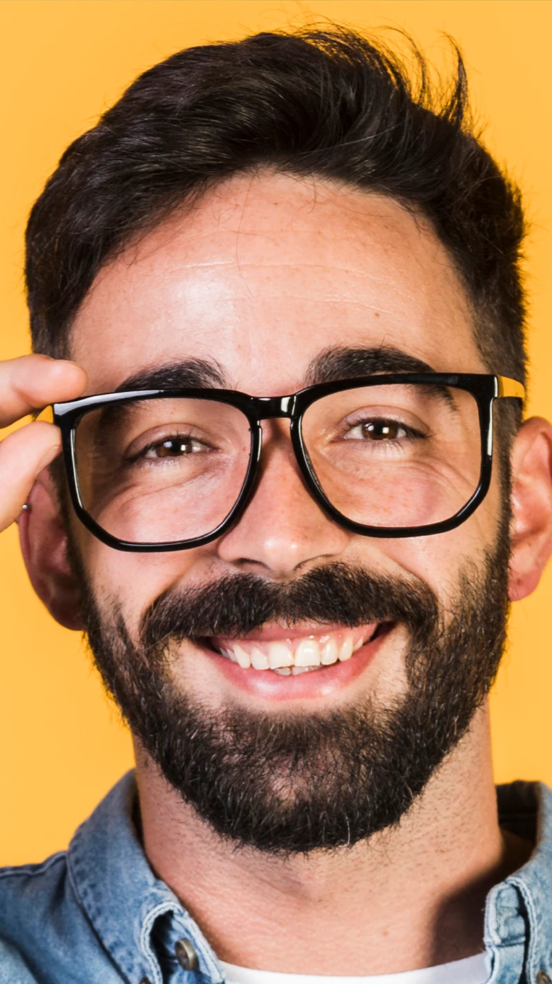 Ein dunkelhaariger Mann mit Bart lacht, während er seine große, schwarze Brille richtet