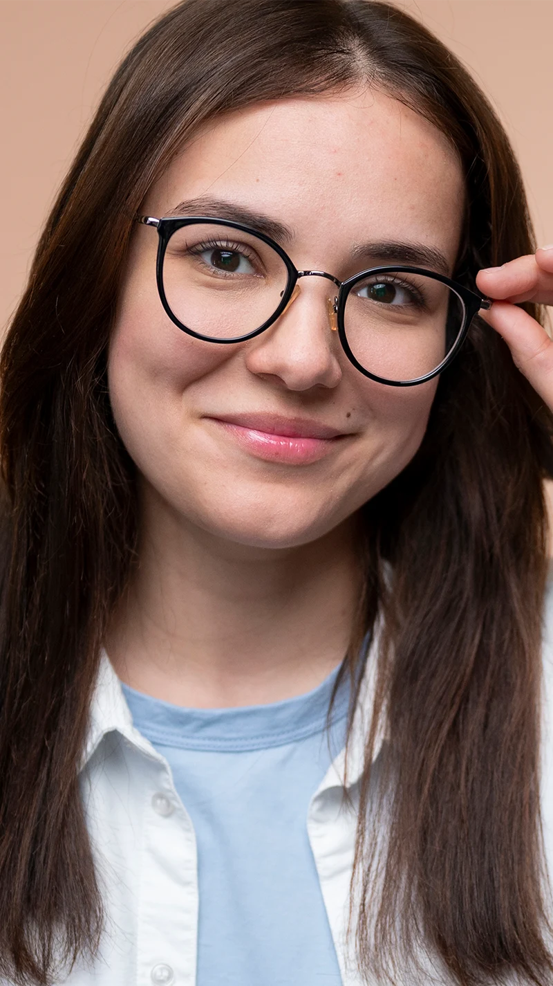 Junge Frau blickt in die Kamera mit einem Lächeln, mit einer Hand hält sie ihre schwarze Brille