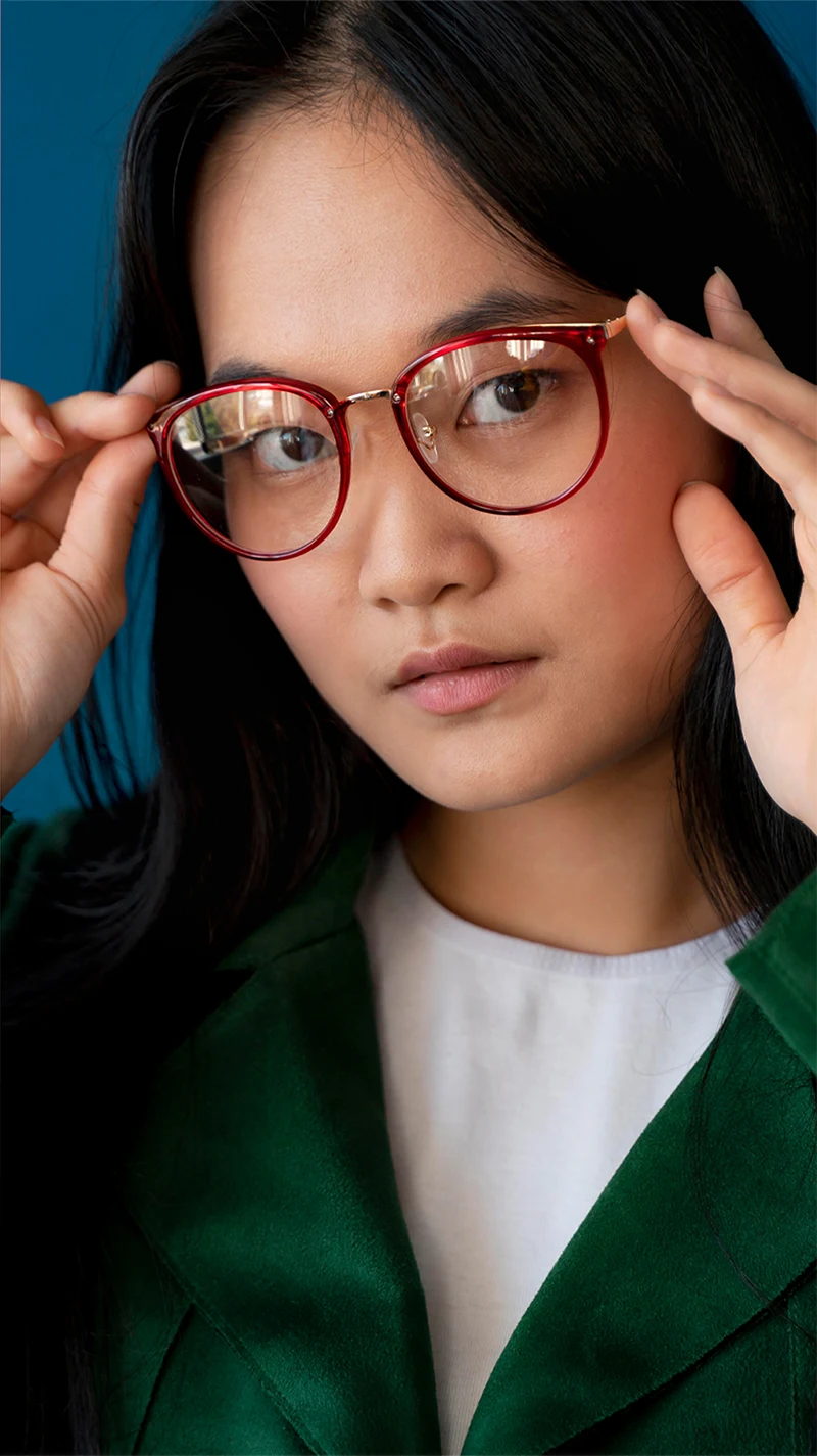 Eine junge Frau ist leicht seitlich zur Kamera gedreht und hält ihre rot gefärbte Brille mit beiden Händen fest