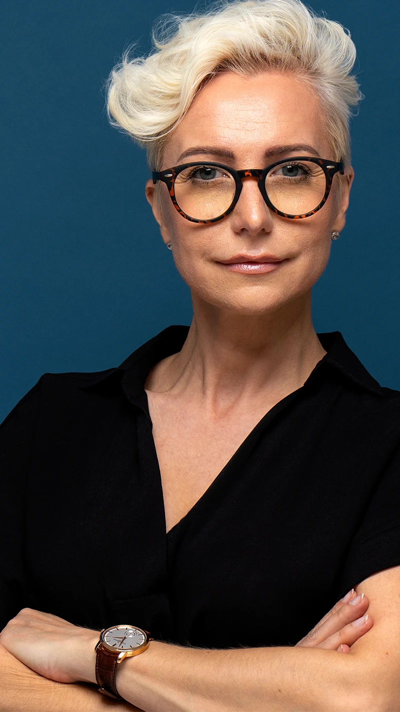 Eine ältere Frau mit kurzem, blondem Haar sieht selbstbewusst in die Kamera. Ihre Brille ist dunkel, mit einer leichten Musterung